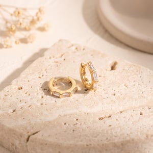 Peut inclure: Une paire de créoles dorées ornées de petites pierres transparentes rectangulaires de taille baguette serties sur le devant de chaque anneau. Les boucles d'oreilles présentent une finition métallique polie et un mécanisme de fermeture à charnière.