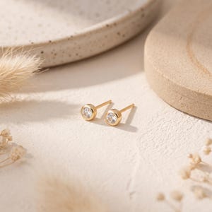May include: A pair of gold stud earrings featuring a single round clear gemstone set in a bezel setting. Each earring has a thin post and a circular gold frame surrounding the central stone, creating a simple and polished appearance for the ears.