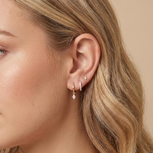May include: A close-up of an ear displaying three gold earrings. The earrings comprise a small hoop with a diamond-shaped pendant, a small hoop with a line of small stones, and a diamond-shaped stud. The earrings are set against a neutral background.