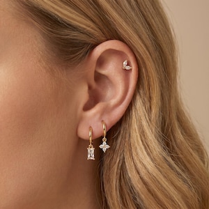 May include: Gold hoop earrings with a rectangular and a four-petal flower-shaped charm. A gold stud earring with two marquise-cut stones is also visible. The jewelry is set against a blurred background.