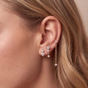 May include: Three gold-toned earrings featuring clear, round-cut stones. One earring is a double-stone stud, another is a vertical drop stud with two stones, and the third is a long chain earring with a single stone at the top and bottom.