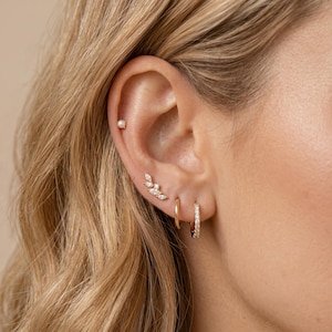 Könnte beinhalten: Ein goldener Ohrschmuck-Stack bestehend aus einem kleinen weißen Perlenstecker am oberen Knorpel, einem marquise-geschliffenen, blattförmigen Kletterohrring in der Mitte und zwei kleinen goldenen Creolen am Ohrläppchen, eine schlicht und eine mit Kristallen.