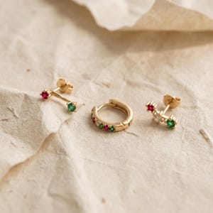 May include: A set of gold jewellery consisting of two stud earrings and one hoop earring. The stud earrings feature a bar design with a red stone and a green stone. The hoop earring is decorated with alternating red, green, and clear stones.