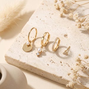 Könnte beinhalten: Ein Set aus vier goldfarbenen Ohrringen, bestehend aus einem Creolen-Ohrring mit flacher runder Scheibe und kleiner Perle, einem Creolen-Ohrring mit hängender Perle und klarem Stein, einem kleinen mit klaren Steinen besetzten Ring und einem Stecker.