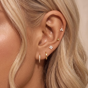 May include: A gold ear stack featuring a small hoop with clear stones, a plain gold hoop, a four-petalled flower stud with clear stones, a small gold ball stud, and a three-stone cluster stud. The jewellery is arranged along the earlobe and cartilage.