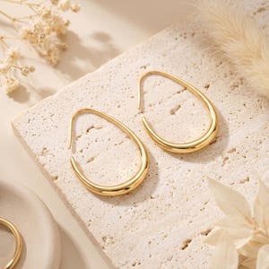 May include: A pair of gold-toned hoop earrings with a smooth, polished finish and an elongated teardrop shape. The earrings feature a continuous, curved metal band that tapers slightly at the top, creating a sleek and uniform appearance.