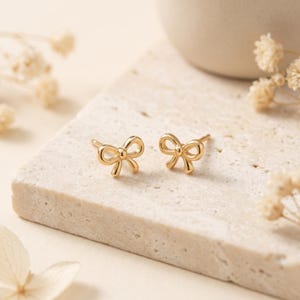 May include: A pair of gold-toned stud earrings shaped like small bows. Each earring features a polished metallic finish with a central knot and two symmetrical loops, designed as a post-style earring for pierced ears with a simple, clean aesthetic.