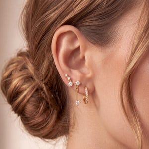 Peut inclure: Trois boucles d'oreilles dorées portées sur une seule oreille. L'une est un petit anneau serti de pierres, une autre est une barre verticale avec deux pierres, et la troisième est un clou incurvé orné de trois pierres de tailles variées.