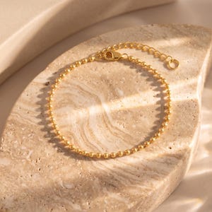 May include: A gold-toned bracelet composed of small, uniform spherical beads linked together in a continuous chain. The bracelet features a lobster claw clasp and an adjustable extension chain with a small circular loop at the very end.