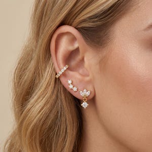 Puede incluir: Juego de joyas para la oreja en tono dorado que incluye un brazalete y tres pendientes. El brazalete está adornado con pequeñas piedras transparentes. Los pendientes incluyen un grupo de tres piedras, un diseño floral y una piedra colgante.
