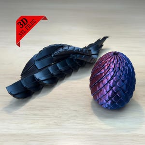 Puede incluir: Una ala y un huevo de dragón impresos en 3D. El ala es negra y segmentada. El huevo es un degradado de morado y azul, con una superficie texturizada y escamosa. La imagen incluye una pancarta roja con el texto "3D STL FILES".