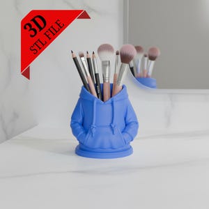 May include: A blue hoodie-shaped desk organizer holding makeup brushes and pencils. The organizer has a hood, pockets, and a round base. A red banner in the corner reads "3D STL FILE". The background includes a mirror and a white countertop.