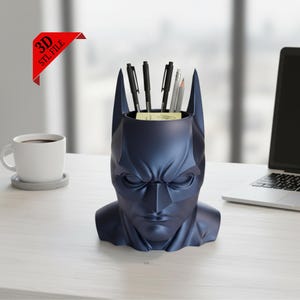 May include: A dark blue Batman head-shaped desk organizer holding pens and pencils. The organizer is on a white desk next to a coffee cup and a laptop. A red banner in the upper left corner reads "3D STL FILE".