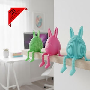 May include: Three colorful bunny figurines in green, pink, and blue sit on a wooden ledge. The egg-shaped bodies have different textures and designs, with articulated legs. The pink bunny has a heart cutout. The image includes the text "3D STL FILES".