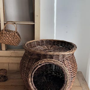 May include: A large, round, brown wicker pet bed with a circular entrance. The bed has a woven design and a matching lid. A smaller woven basket and a woven handbag are also visible.