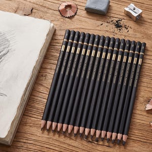 May include: A set of sixteen black drawing sketch pencils arranged in a row, each labeled with its specific grade ranging from 4H to 12B. The pencils feature gold lettering and are positioned next to a sketchbook, a gray eraser, and a metal sharpener.