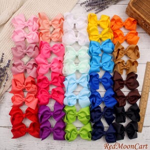 May include: A collection of thirty-two solid-colored hair bows arranged in a grid, featuring shades of pink, orange, yellow, white, blue, brown, red, purple, green, and black. A wooden ruler is visible on the right side of the image.