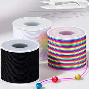 May include: Three spools of cord in black, white, and rainbow colors. A pair of white scissors and a needle threader are in the background. A string of colorful beads is attached to the rainbow cord. The spools are on a white surface.
