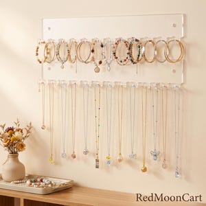 May include: A clear acrylic wall-mounted jewelry organizer featuring two horizontal rows of hooks. The top row holds various bracelets, while the bottom row displays multiple necklaces with diverse pendants. The text RedMoonCart is visible.