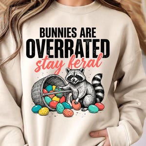 May include: A beige sweatshirt with the text "Bunnies Are Overrated Stay Feral". The graphic features a raccoon with a basket of colorful Easter eggs. The raccoon is sitting next to the basket, with some eggs scattered around.