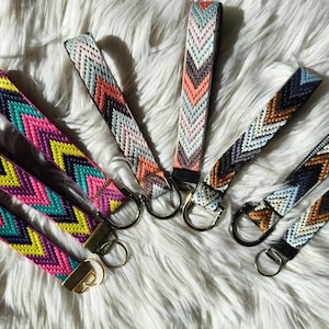 May include: A selection of woven wrist keychains with chevron patterns in various colours. The keychains display pink, yellow, teal, orange, and brown hues. Each keychain has a metal ring and a fabric loop for convenient carrying.