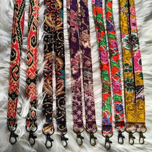 May include: A collection of long fabric lanyards featuring various colourful patterns including red, black, yellow, and purple floral and geometric designs. Each strap is finished with a metal swivel lobster clasp at the bottom end.