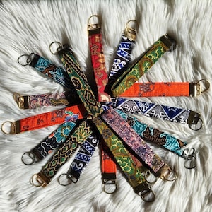 Puede incluir: Una colección de llaveros de tela coloridos con varios diseños, incluyendo florales y geométricos. Cada llavero presenta un anillo metálico y un lazo de tela en tonos rojos, naranjas, verdes, azules y morados.