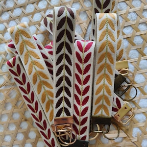 May include: A collection of fabric wristlet keychains featuring a woven leaf pattern in shades of mustard yellow, deep red, and dark brown on a cream background. Each strap is secured with a metal hardware clasp and a circular split ring.