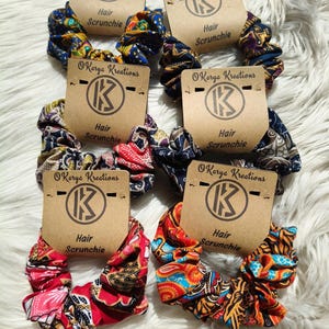 May include: Six fabric hair scrunchies with various colourful patterns, each attached to a tan cardboard display card labelled Karye Kreations and Hair Scrunchie. The scrunchies feature intricate designs in shades of red, blue, yellow, and black.