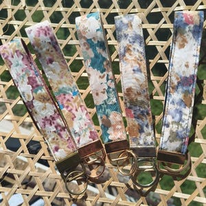 May include: Five fabric wristlet keychains featuring various floral and abstract patterns in shades of pink, teal, blue, and tan. Each strap is secured with a metallic clasp and includes a circular metal ring for attaching keys or accessories.