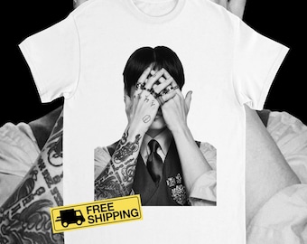 May include: White t-shirt featuring a black and white image of a person with tattoos and rings covering their face. The person is wearing a vest and tie. The image is a close-up of the person's upper body.
