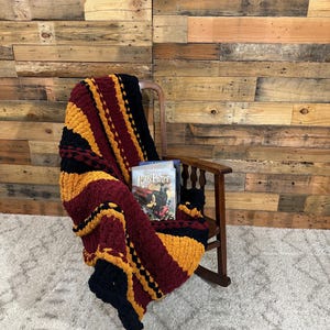 May include: A chunky knit blanket in burgundy, gold, and black draped over a wooden rocking chair. The blanket has a striped pattern. A copy of "Harry Potter" is resting on the chair. The background is a wooden wall and a white rug.