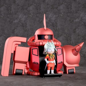 Może przedstawiać: Czerwona zabawka robota z małą figurką pilota w kokpicie. Robot ma zaokrągloną głowę z płetwą, duże ciało i detal w kształcie rogu. Pilot nosi hełm i pomarańczowy mundur.