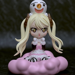 May include: A chibi-style figurine of a blonde-haired character with brown horns, wearing a pink and white dress, and black gloves. A smiling snowman sits atop the character's head. The figurine rests on a pink cloud.