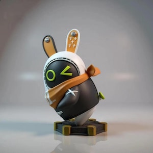 May include: A stylized rabbit figurine with a white head, black body, and orange accents. The figurine has a scarf, a green button, and a base with gold-colored edges. The rabbit's head features a black visor with a green circle and a yellow chevron.
