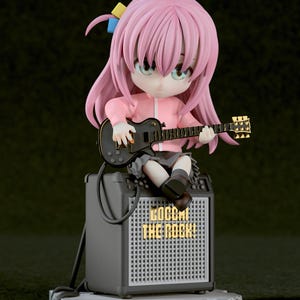 Puede incluir: Figura de un personaje con pelo rosa, que lleva una chaqueta rosa y una falda gris, tocando una guitarra negra. La figura está sentada en un amplificador gris con el texto "BOCCHI THE ROCK!".