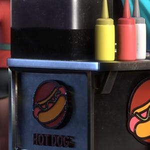 May include: A black hot dog cart with a red, yellow, and white condiment bottles. The cart has a circular logo with a hot dog and the words "HOT DOG" in red letters.