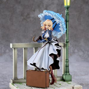 Può includere: Statuetta di una donna bionda con un vestito blu e bianco, che tiene un ombrello blu. Una valigia marrone è vicina. Un lampione verde è accanto. La statuetta è su una base di pietra.