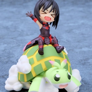 May include: A figurine of a cartoon character with black hair and red and black armor, sitting atop a green and yellow turtle-like creature with white accents. The turtle has a smiling face and pink accents.