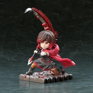 Puede incluir: Figura de un personaje de la serie RWBY, posando con una gran guadaña roja. La figura lleva un atuendo negro y rojo con una capa roja. La base es una plataforma gris con detalles rojos.