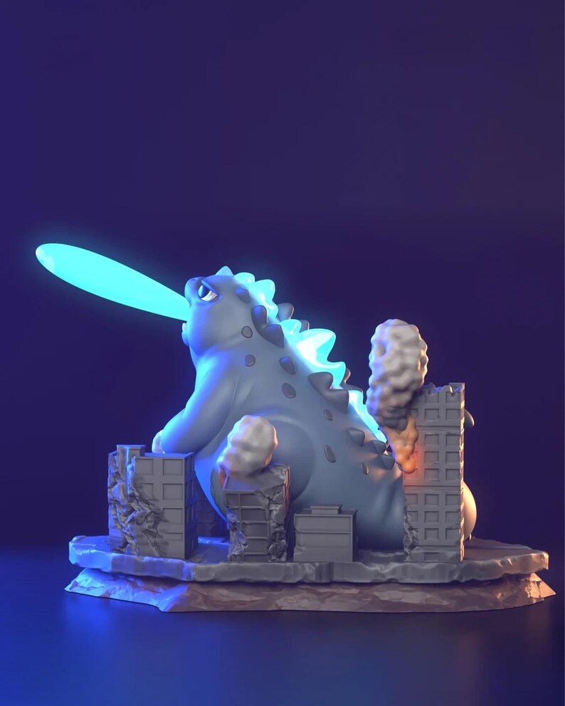 May include: A grey Godzilla-style figurine with a blue dorsal fin and a blue beam coming from its mouth. The figurine is destroying miniature grey buildings, with smoke and flames visible. The base is a textured, rocky surface.