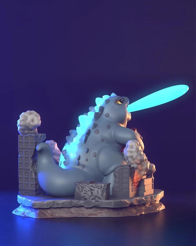 May include: A light blue Godzilla figurine with a glowing blue atomic breath. The figurine is destroying miniature buildings, with smoke and rubble details. The figure has a textured back and is set on a stone base.