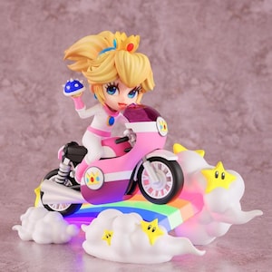 Peut inclure: Figurine de la Princesse Peach sur une moto rose et blanche. La figurine est posée sur une base arc-en-ciel avec des nuages blancs et des étoiles jaunes. Peach porte une tenue blanche et tient un champignon bleu.