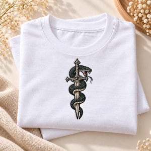 May include: A white t-shirt featuring a central graphic print of a dark green snake coiled around a silver-toned sword. The snake has visible scales, an open mouth with fangs, and a forked tongue. The sword has a detailed hilt and a pointed blade.
