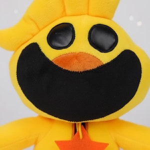 Pode incluir: Um brinquedo de pelúcia amarelo com olhos e boca pretos e um nariz laranja. O brinquedo tem uma etiqueta laranja em forma de estrela e um tufo de cabelo amarelo. O brinquedo foi projetado para se parecer com um personagem de desenho animado.