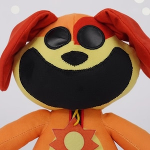 Pode incluir: Um cão de pelúcia com um rosto sorridente. O cão tem uma cabeça amarela e laranja com olhos pretos grandes e uma boca preta larga. As orelhas são vermelhas e o corpo é laranja com um emblema amarelo em forma de sol.