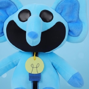 Puede incluir: Un peluche de elefante azul claro con ojos negros y boca negra. El elefante tiene orejas grandes y una trompa larga. Una etiqueta amarilla con un diseño de bombilla cuelga de su cuello.