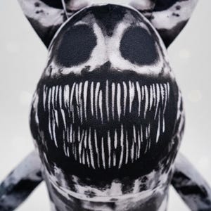 May include: A black and white plush toy with a distinctive design. The toy has a large, toothy grin and large eyes. The plush has a soft, textured appearance and is set against a blurred background.