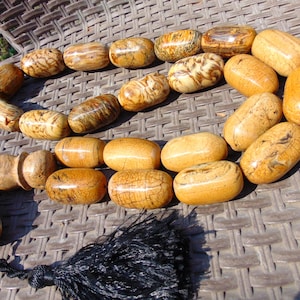 May include: A prayer bead strand featuring polished, barrel-shaped beads with a marbled tan and brown wood-grain pattern. The strand includes a larger, textured end bead and a long, black silk tassel attached to the base of the prayer beads.