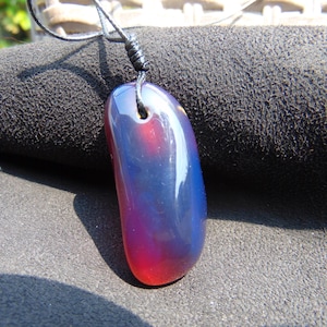 Puede incluir: Un colgante oblongo pulido con un degradado de colores, que transita del rojo al azul. El colgante está suspendido de un cordón negro. La piedra preciosa tiene una superficie lisa y brillante.
